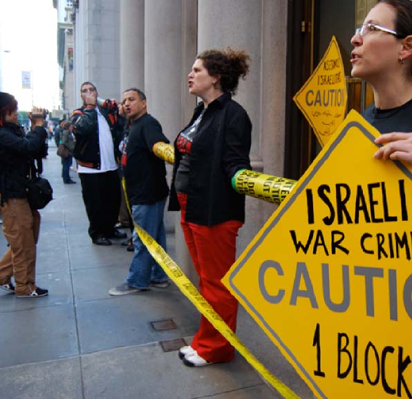 IJAN organizers holding caution-sign placards reading 'Israeli War Crimes' at a street demonstration.